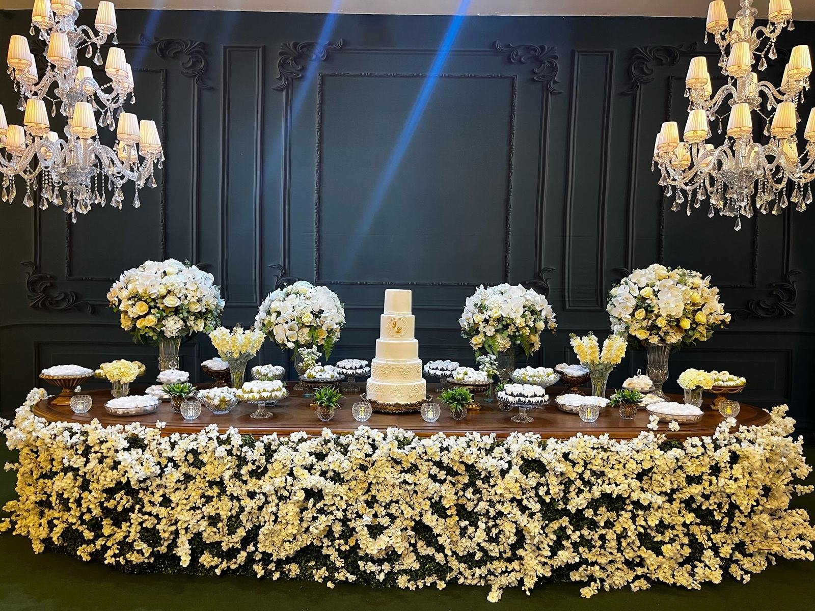 Mesa do bolo coberta de flores brancas diante de uma parede em boiserie verde, entre dois lustres de cristal