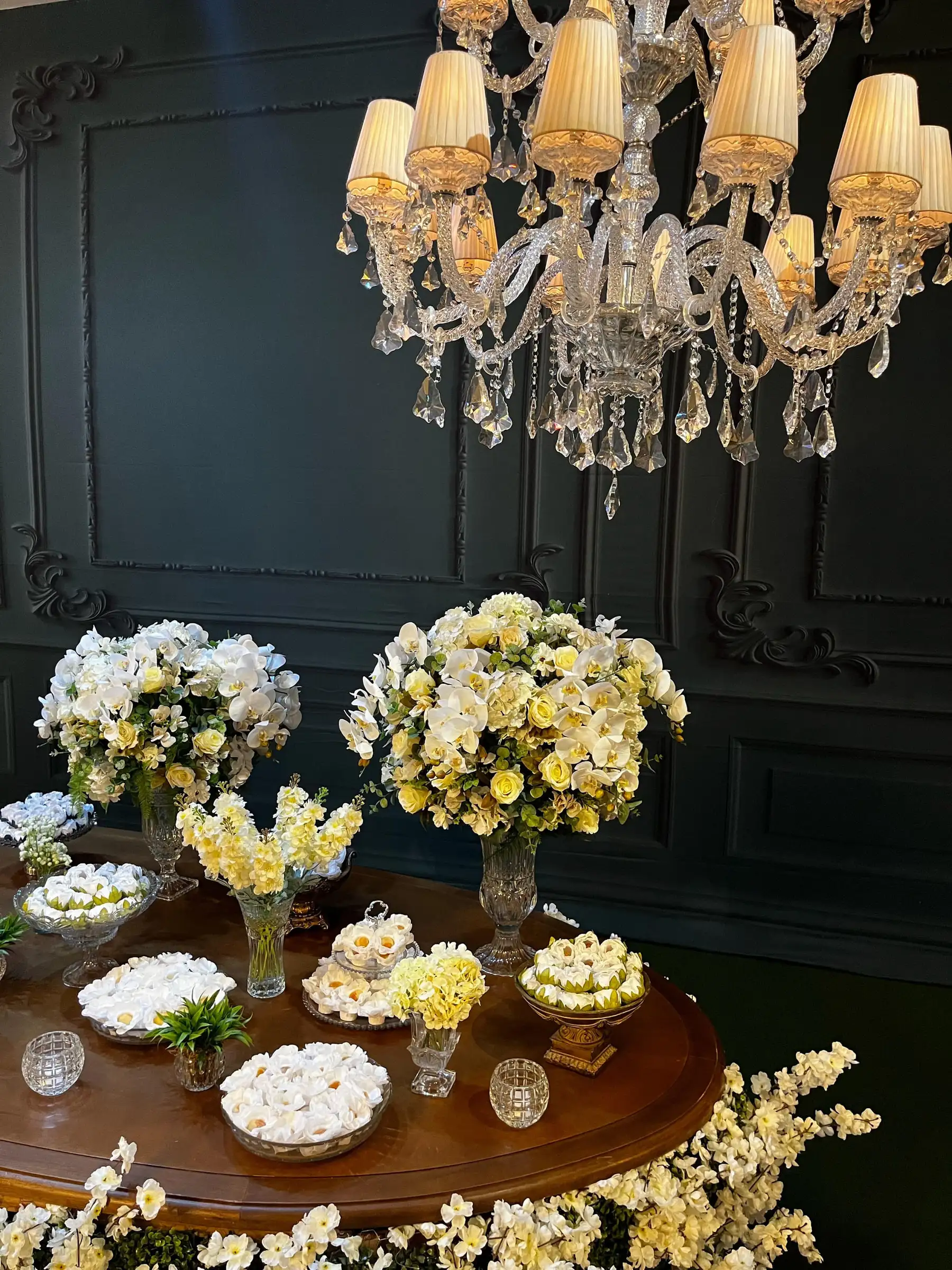 Lustre de cristal sobre mesa redonda de doces com arranjos de flores brancas e amarelas
