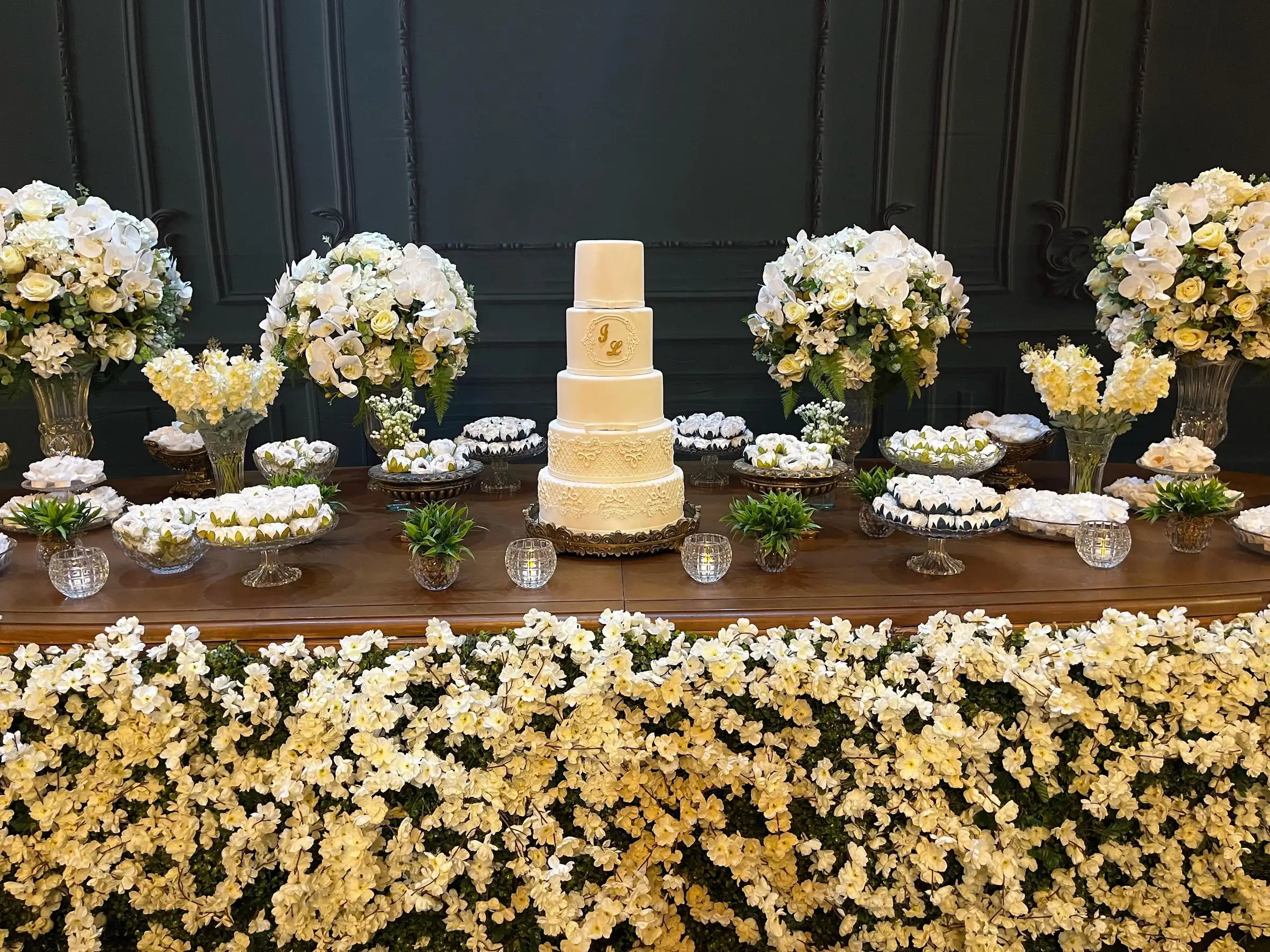 Mesa do bolo de casamento coberta de flores brancas e amarelas, com arranjos altos diante da parede verde